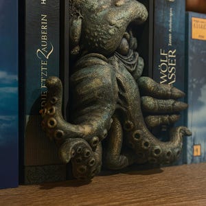 May include: A bronze-colored octopus bookend sculpture. The tentacles wrap around the books, with the body of the octopus emerging from the center. The bookend is placed between two books on a wooden shelf. The books have titles in German.