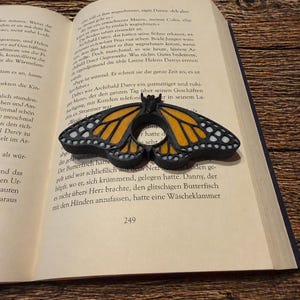 Könnte beinhalten: Ein schwarzer und orangefarbener Schmetterlingsring, der einem Monarchfalter ähnelt, liegt auf einem offenen Buch. Der Ring hat detaillierte Flügelmuster mit schwarzen und weißen Akzenten. Die Seiten des Buches sind sichtbar, zusammen mit Text und der Nummer 249.