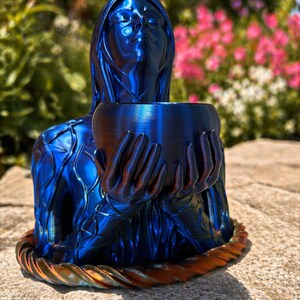 Könnte beinhalten: Eine blaue Skulptur einer Frau, die eine Schale hält. Die Skulptur hat eine metallische Oberfläche mit Blau- und Bronzetönen. Das Gesicht der Frau ist nach oben geneigt, und die Schale wird vor ihr gehalten. Der Sockel der Skulptur hat ein gedrehtes Seildesign.