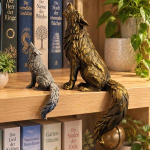 Könnte beinhalten: Zwei Wolfsfiguren in heulender Pose, eine kleinere in verwitterter Silberoptik und eine größere in metallischem Bronze-Finish, beide mit detaillierter Fellstruktur und langen Schwänzen, die über den Rand eines Holzregals hängen.