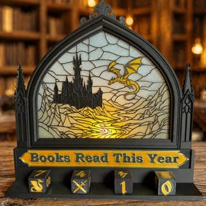 Könnte beinhalten: Schwarz-goldene Anzeige im Glasmalerei-Stil mit Burg- und Drachendesign. Die Anzeige lautet "Books Read This Year" und hat Würfel zur Verfolgung des Lesefortschritts. Der Hintergrund ist eine verschwommene Bibliothek.