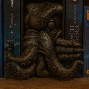 May include: A bronze-colored octopus sculpture, mounted between two dark blue books. The sculpture has detailed tentacles and a textured surface. The books have text in German. The sculpture is on a wooden shelf.