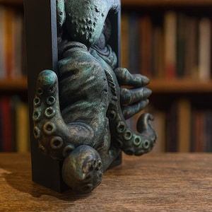 May include: A detailed sculpture of a creature with tentacles, emerging from a black rectangular frame. The sculpture has a weathered, greenish-bronze finish. The background is a blurred bookshelf.