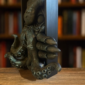 May include: A bronze-colored octopus sculpture emerging from a black rectangular frame. The sculpture has detailed tentacles and a textured surface, resting on a wooden surface. The background features a blurred bookshelf.