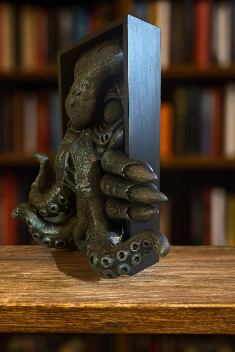 May include: A bronze-colored octopus sculpture emerging from a black rectangular frame. The sculpture has detailed tentacles and a textured surface, resting on a wooden surface. The background features a blurred bookshelf.