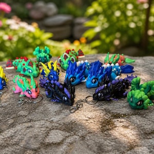 Könnte beinhalten: Eine Sammlung farbenfroher Drachen-Schlüsselanhänger in verschiedenen Blau-, Grün-, Rosa- und Lilatönen. Jeder Drache hat ein einzigartiges Design und ist an einem silbernen Schlüsselanhänger befestigt. Die Schlüsselanhänger liegen auf einer strukturierten Steinoberfläche mit unscharfem grünem Hintergrund.