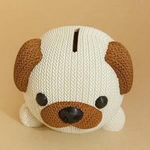 May include: A ceramic pug-shaped piggy bank. The bank is white with brown accents on the ears, snout, and around the eyes. It has a slot on top for inserting coins and is a decorative item.
