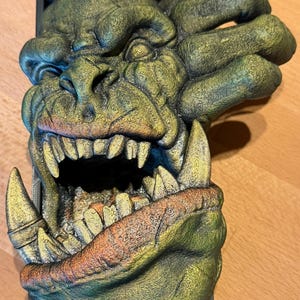 以下が含まれることがあります： 鋭い歯と開いた口を持つ、緑と金のモンスターの顔の精巧な彫刻。テクスチャ加工された彫刻は、黒い長方形のベースに取り付けられています。モンスターは、顕著な眉、鼻、大きな爪のような手を持っています。