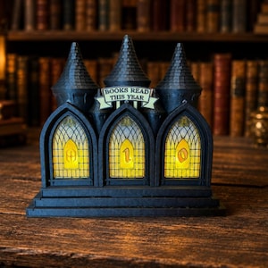 Könnte beinhalten: Ein schwarzer und grauer Bücherzähler in Burgform mit drei gewölbten Fenstern, die die Zahl "10" anzeigen. Der Text "BOOKS READ THIS YEAR" befindet sich über den Fenstern. Der Zähler steht auf einer Holzoberfläche, mit einem Bibliotheks-Hintergrund.