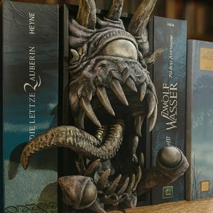 Könnte beinhalten: Ein dunkel strukturierter Buchständer, der ein skulptiertes Monster mit einem großen zentralen Auge, scharfen Zähnen und einer herausragenden Zunge zeigt. Die Kreatur befindet sich in einem rechteckigen Rahmen zwischen zwei Büchern mit Text.
