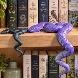 Könnte beinhalten: Zwei bewegliche Schlangenfiguren auf einem Holzregal. Eine Schlange ist dunkelblau mit gelben Augen, die andere ist eine größere, leuchtend violette Schlange mit hellblauen Augen. Beide haben detaillierte, strukturierte Schuppen und sind gewunden.