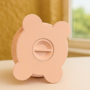 May include: A light pink piggy bank shaped like a bear. The bank has four rounded ears and a slot for inserting coins. The coin slot is located on the front of the bank. The bank is made of a textured material.