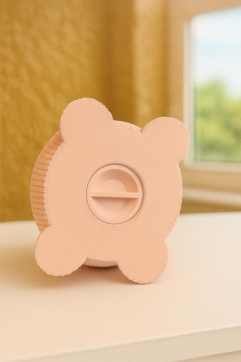 May include: A light pink piggy bank shaped like a bear. The bank has four rounded ears and a slot for inserting coins. The coin slot is located on the front of the bank. The bank is made of a textured material.