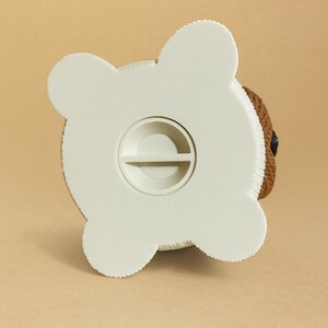 May include: A white and brown teddy bear-shaped piggy bank. The bottom of the bank is white with a coin slot. The bear's head is brown with a black eye. The bank is on a tan background.
