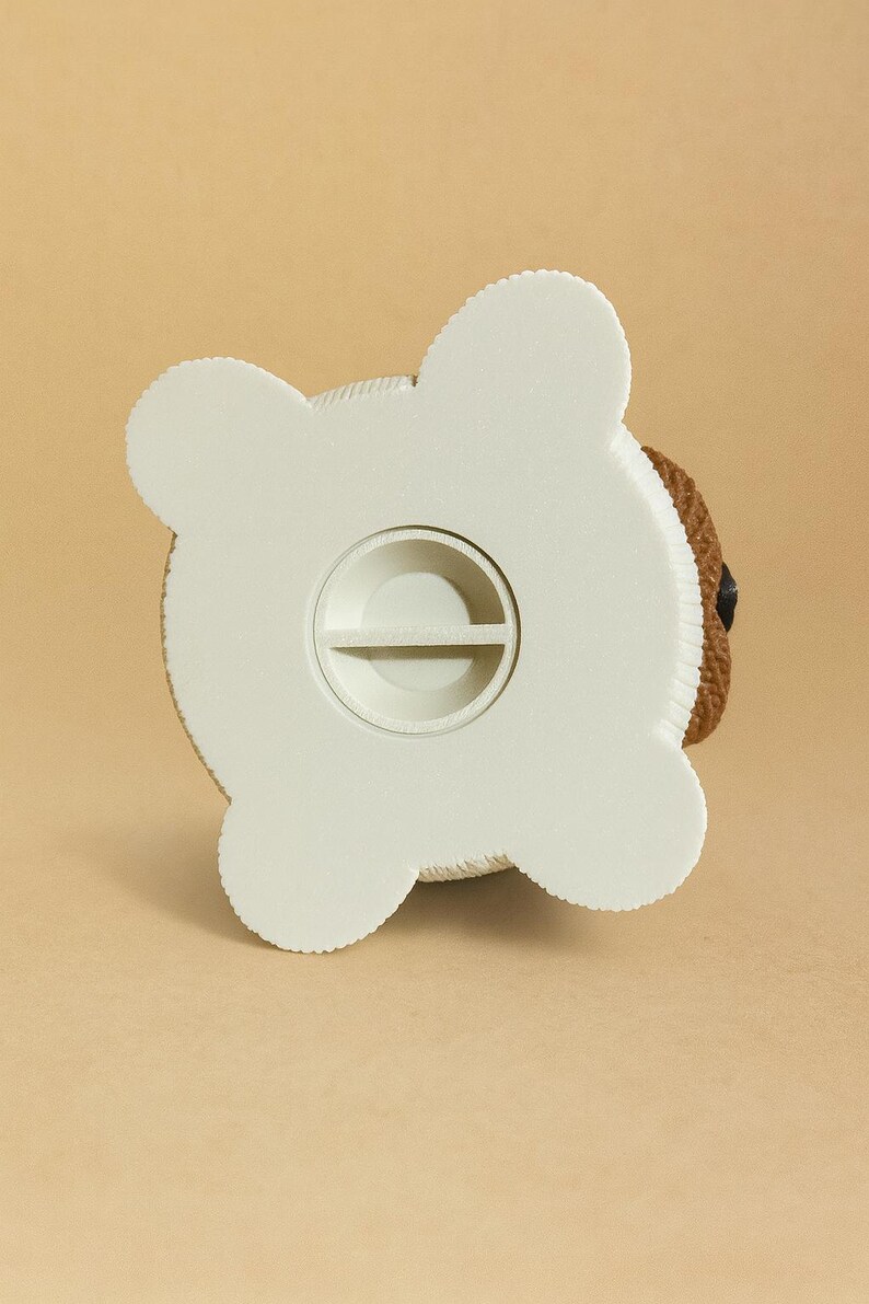 May include: A white and brown teddy bear-shaped piggy bank. The bottom of the bank is white with a coin slot. The bear's head is brown with a black eye. The bank is on a tan background.