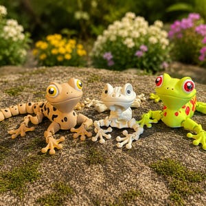 Könnte beinhalten: Drei Spielzeugfiguren von Reptilien und Amphibien werden im Freien gezeigt. Links ist eine hellbraune Eidechse mit schwarzen Flecken und einem segmentierten Schwanz. In der Mitte befindet sich ein weißer Skelettfrosch. Rechts ist ein grüner Frosch mit roten Flecken.