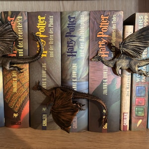 Op de afbeelding: Drie bronskleurige drakenbeeldjes met gedetailleerde vleugels en lange staarten, geplaatst op de ruggen van een rij diverse boeken. De draken zijn in een verspringende formatie gerangschikt, met twee draken bovenaan en één draak eronder.