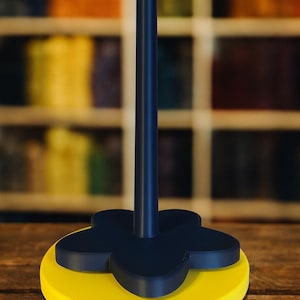 Op de afbeelding: Een decoratieve standaard met een gele basis en een donkerblauwe bloemvormige bovenkant. Een donkerblauwe paal komt uit het midden. De standaard staat op een houten oppervlak met een vage achtergrond van planken.