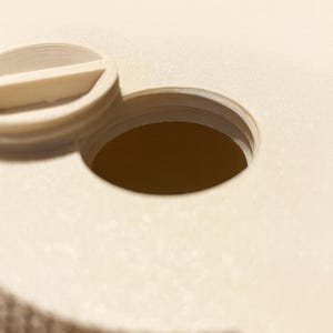 May include: Close-up of a beige plastic object with a circular opening. The object has a textured surface and a small, curved, raised section. The opening is dark brown, and the overall design is simple and functional.