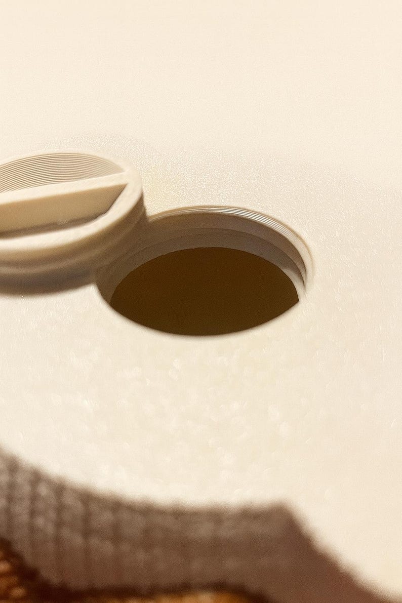 May include: Close-up of a beige plastic object with a circular opening. The object has a textured surface and a small, curved, raised section. The opening is dark brown, and the overall design is simple and functional.