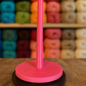 Może przedstawiać: Różowy motowidło do przędzy z czarną podstawą, przeznaczone do trzymania przędzy. Motowidło ma wysokie, centralne wrzeciono i okrągłą podstawę. Tło przedstawia półki wypełnione kolorowymi motkami przędzy.