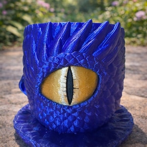 Könnte beinhalten: Blauer Drachen-Pflanztopf mit strukturierter Oberfläche. Der Pflanztopf hat ein großes, gesticktes goldenes Auge mit schwarzer Pupille. Das Design umfasst Schuppen und einen stacheligen Rand.
