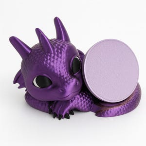 Op de afbeelding: Een paars drakenbeeldje met grote zwarte ogen en een ronde, lichtpaarse basis. De draak zit in een rustpositie, met zijn hoofd omhoog en zijn staart opgerold. Het beeldje is gemaakt van een textuurrijk materiaal.