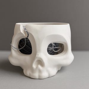 Op de afbeelding: Een witte keramische schedelvormige plantenbak met grote oogkassen en een scheur aan de bovenkant. De binnenkant bevat een bolletje grijs garen. De plantenbak staat op een grijze ondergrond.