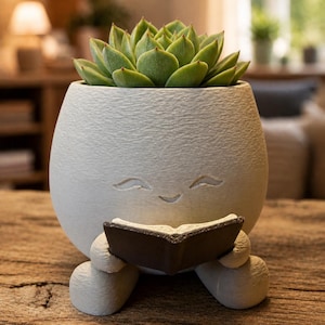 Può includere: Un'eccentrica fioriera in ceramica grigio chiaro a forma di personaggio sorridente, con un piccolo libro in mano. La fioriera è riempita con una succulenta verde vibrante. La fioriera poggia su una superficie di legno.