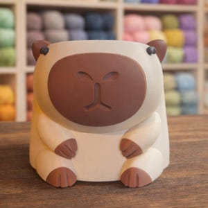 May include: A cream-colored, bear-shaped planter with brown accents. The planter has a rounded body, a brown face with a simple nose and eyes, and small brown ears. The planter is sitting on a wooden surface, with shelves of colorful yarn in the background.