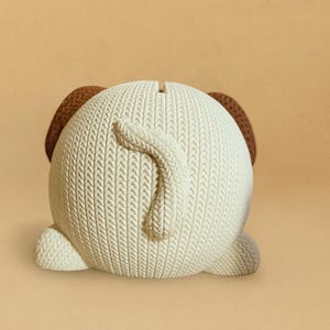 May include: A ceramic dog-shaped bank with a textured, off-white body and brown ears. The dog's tail curls upwards. The bank has a slot on top for inserting coins. The design resembles a knitted sweater.
