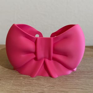 Könnte beinhalten: Ein leuchtend rosa, schleifenförmiger Behälter. Der Behälter hat eine abgerundete Form mit einem zentralen Knoten und zwei Schlaufen, die einer großen Schleife ähneln. Der Behälter besteht aus einem glatten, festen Material und steht auf einer Holzoberfläche.