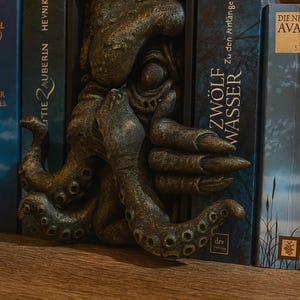 May include: A bronze-colored octopus bookend with detailed tentacles and a textured surface. The bookend is placed between two books with text on the spines, including "ZW&Ouml;LF WASSER" and "TIE ZAUBERIN HEYNIK".