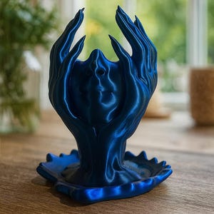 Op de afbeelding: Een donkerblauwe decoratieve sculptuur met een gezicht en omhoog reikende handen. De sculptuur is gemaakt van een glanzend materiaal en staat op een geschulpte basis. De achtergrond is wazig.