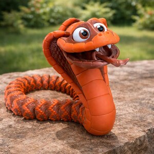 Op de afbeelding: Een grillig oranje en bruin cobra-beeld met een wijd open mond en uitgestoken tong. Het lichaam van de slang is opgerold en het beeld staat op een stenen oppervlak met een vage groene achtergrond.