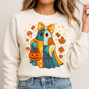 Puede incluir: Sudadera color crema con un personaje de fantasma de patchwork sosteniendo un cubo de calabaza de Halloween. El fantasma tiene un gran lazo y está rodeado de gráficos temáticos de otoño como hojas, setas y estrellas. El diseño es colorido y caprichoso.