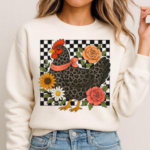 May include: Cream-coloured sweatshirt with a graphic of a chicken with a leopard print pattern, a red comb, and a pink scarf. The chicken is surrounded by flowers, including sunflowers and roses, set against a black and white chequered background.