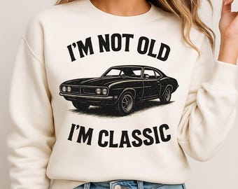 Könnte beinhalten: Cremefarbenes Sweatshirt mit dem Text "I'M NOT OLD I'M CLASSIC" über und unter einer schwarzen Illustration eines Oldtimers. Das Auto ist ein klassisches Coupé-Design mit detaillierten Scheinwerfern und einer schlanken Karosserie.