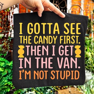 Könnte beinhalten: Schwarze Tragetasche mit dem Text "I GOTTA SEE THE CANDY FIRST. THEN I GET IN THE VAN. I'M NOT STUPID" in Gelb, Rosa und Orange. Die Tasche wird von einer Hand gehalten.