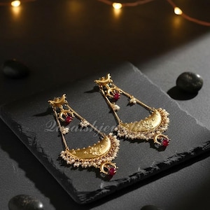 Könnte beinhalten: Goldfarbene Halbmond-Ohrringe mit floralen Details, kleinen roten Edelsteinen und hängenden Perlen. Die Ohrringe werden auf einer dunklen Oberfläche mit dekorativen Lichtern im Hintergrund präsentiert.