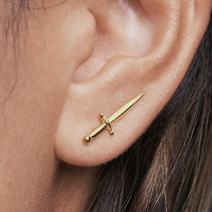 May include: A gold-toned stud earring shaped like a small sword. The earring features a pointed blade, a central crossguard, and a rounded pommel at the base. The piece is positioned horizontally across the earlobe, showcasing a polished metallic finish.