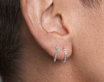 Puede incluir: Un par de pendientes de aro pequeños y finos de plata colocados en el lóbulo de la oreja. Los aros presentan una superficie delicadamente texturizada con un patrón repetitivo en el borde exterior, creando un brillo metálico sutil.