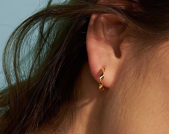 Op de afbeelding: Een kleine gouden oorring met een gedraaide, spiraalvormige textuur. De oorring is cirkelvormig en heeft een gepolijste, reflecterende metalen afwerking die het licht opvangt, waardoor een subtiel, herhalend patroon langs de band ontstaat.