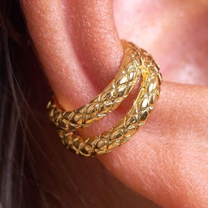 May include: A gold ear cuff featuring a double-band design with a textured, quilted diamond pattern across the surface. The jewelry piece wraps around the outer edge of the ear cartilage, displaying a polished metallic finish and a repeating geometric motif.