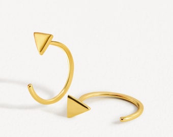 Peut inclure: Une paire de boucles d'oreilles créoles dorées, chacune dotée d'une petite breloque triangulaire pleine fixée au fil incurvé. Les créoles sont ouvertes et le métal présente une finition lisse et polie qui reflète la lumière, créant un aspect métallique brillant.