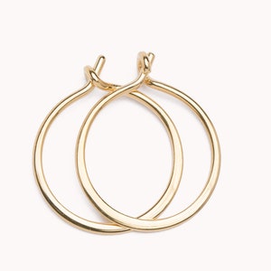 Könnte beinhalten: Ein Paar polierte goldfarbene Creolen mit kreisförmigem Design und Klappverschluss. Die Ohrringe haben eine glatte, reflektierende metallische Oberfläche und eine einfache, durchgehende runde Form, die sich am oberen Verschluss leicht überlappt.