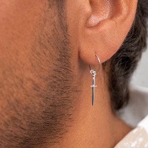 May include: A silver earring in the shape of a small sword is worn in the ear. The earring hangs from a thin silver hoop. The person is wearing a white and tan plaid shirt.