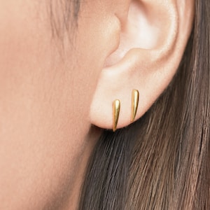 May include: Two small, polished gold-tone stud earrings shaped like elongated, tapered spikes. The earrings are positioned vertically in the earlobe, one slightly higher than the other, featuring a smooth, reflective metallic surface finish.