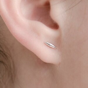 May include: A small, polished silver-tone stud earring shaped like a smooth, elongated grain or rice kernel. The earring is positioned in the lower lobe of an ear, featuring a metallic, reflective surface with a tapered, rounded design.