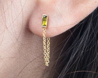 May include: A gold stud earring featuring a rectangular, olive-green baguette-cut gemstone. A delicate gold chain hangs from the base of the gemstone setting, dangling downward from the earlobe to create a subtle, layered, and dangling effect.